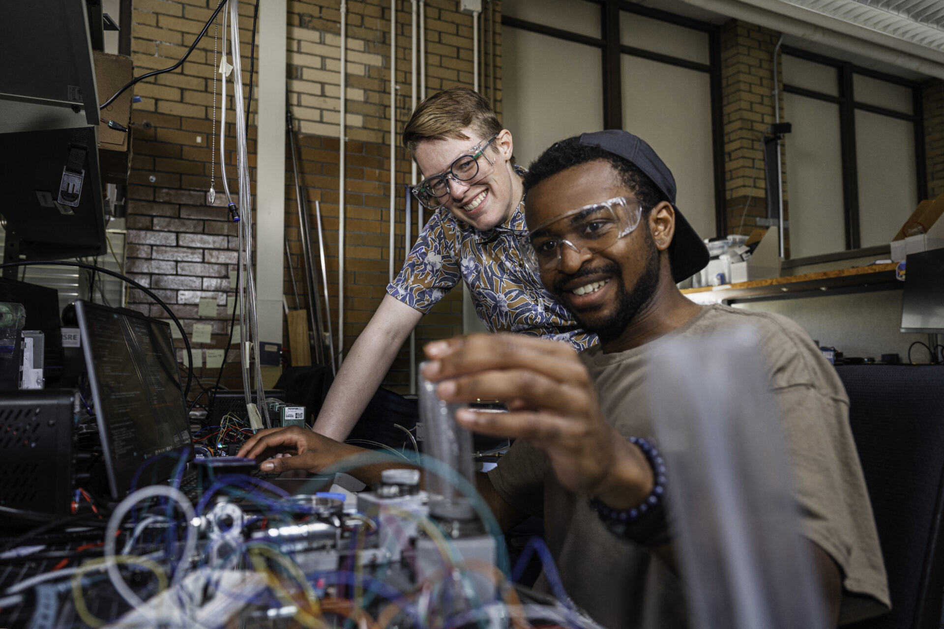Two individuals wearing safety goggles collaborate on a complex electronic setup in a workshop environment.