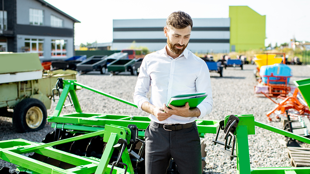 A salesman standing near the plow at the outdoor ground of the shop with new agricultural machinery
