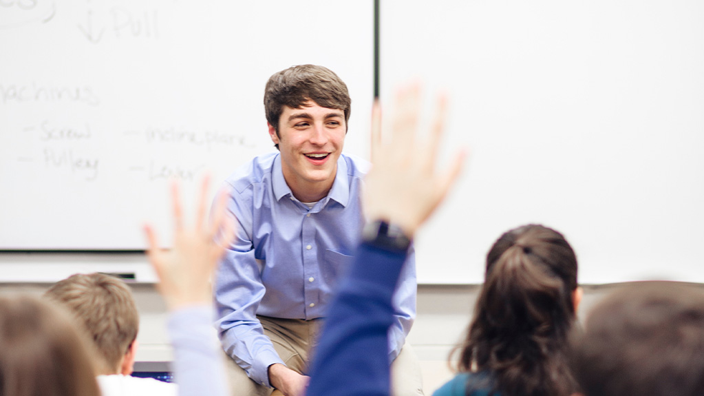 Student teacher smiling as young children raise their hand.