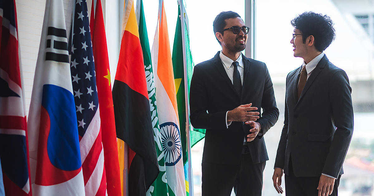 Two businessmen greet each other next to a display of global flags