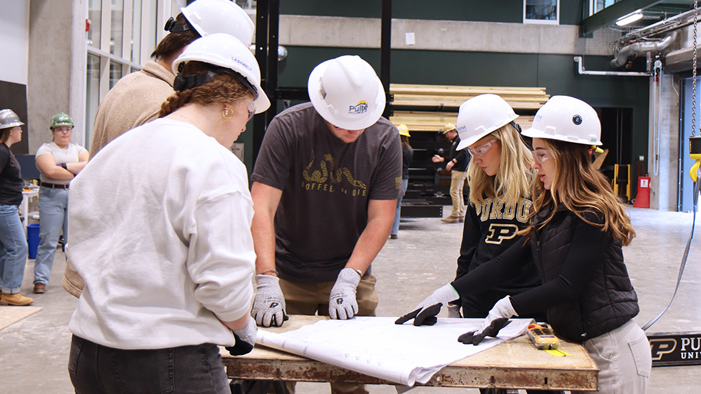 Students wear protective gear as they study blueprints standing around a table