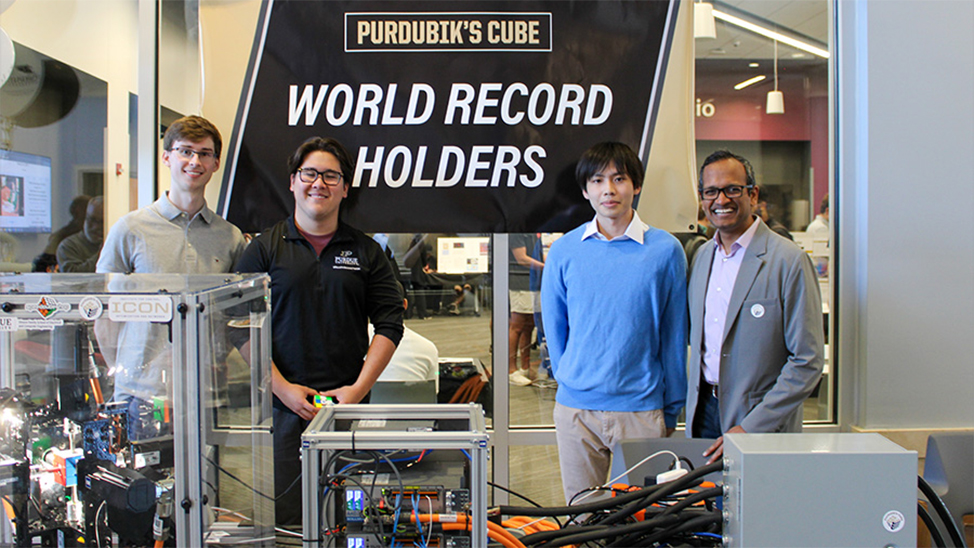 The Purdubik's Cube and their department head stand behind the robot with a World Record Holders flag behind them.