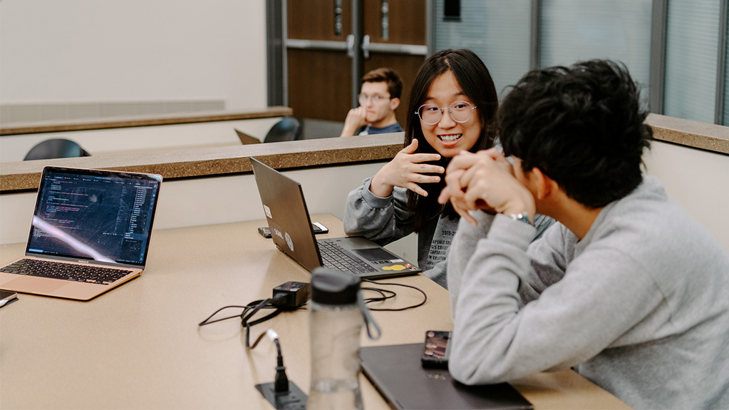 two students talking at a table in front of lap tops