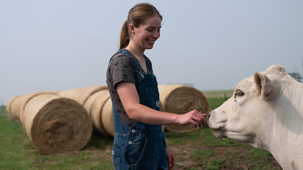 Emma Bailey in denim overalls smiles as she gently reaches out to a white cow in a grassy field, with large round hay bales lined up in the background.