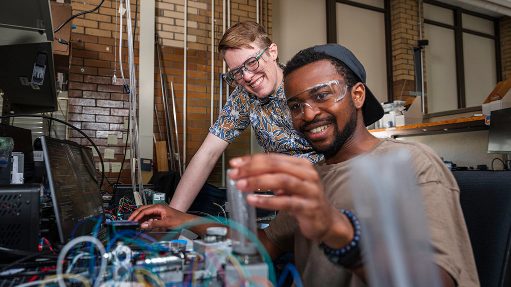 Two individuals wearing safety goggles collaborate on a complex electronic setup in a workshop environment.