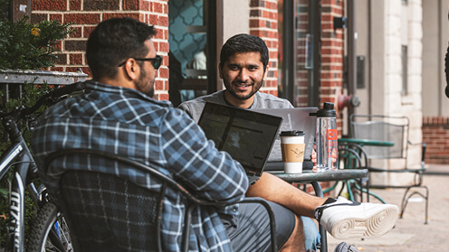 Students talking at outside table with laptop and coffee.