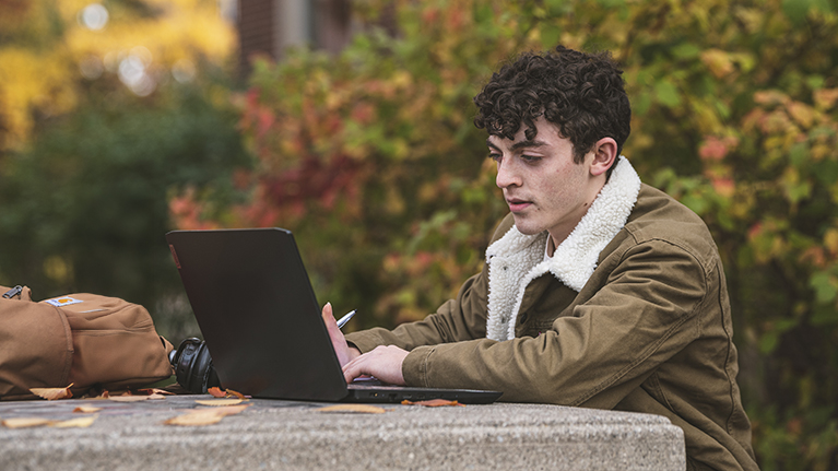A student works on his laptop at a table outside with fall leaves in the background