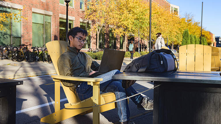 A student sits outside working on his laptop at a firepit station on Third Street