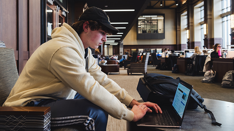 A student sits in WALC working on his laptop with students sitting at workstations in the background