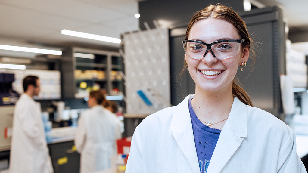 A female student smiles standing in a laboratory wearing protective gear