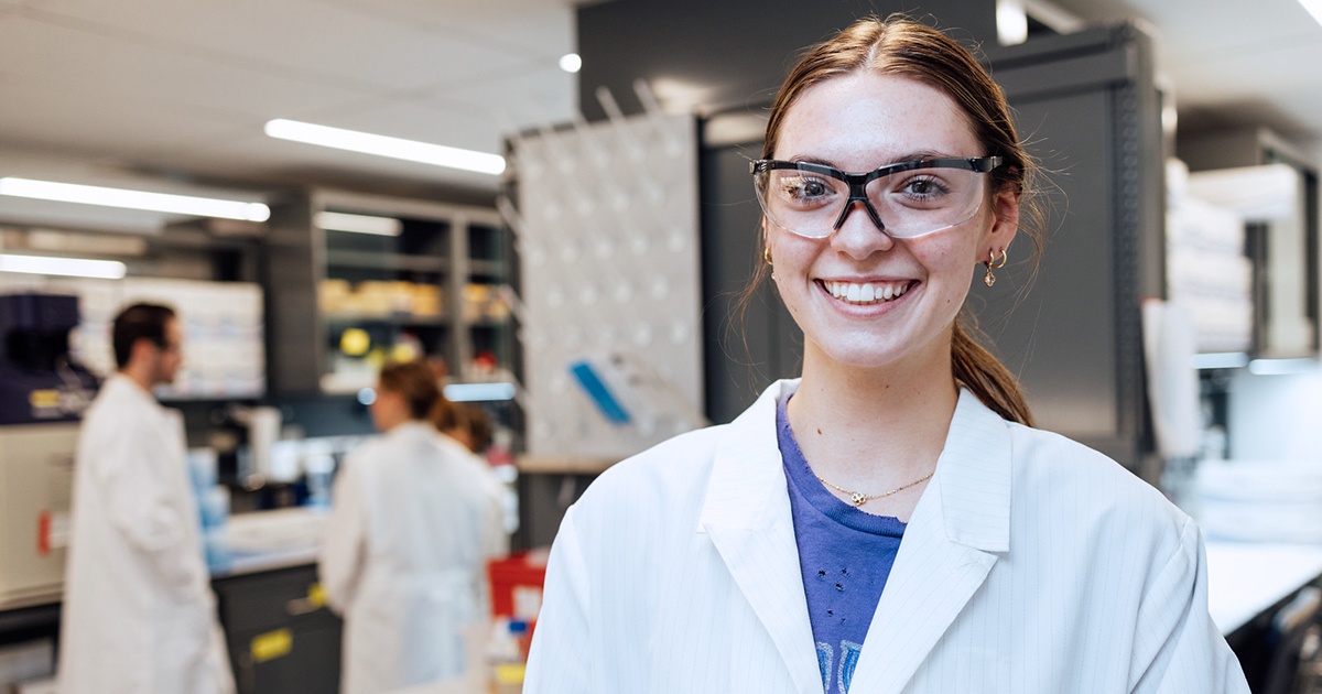 A female student smiles standing in a laboratory wearing protective gear