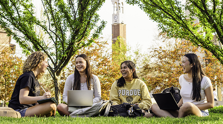 Four students sit together laughing on the Engineering Mall with the Purdue Bell Tower in the background.