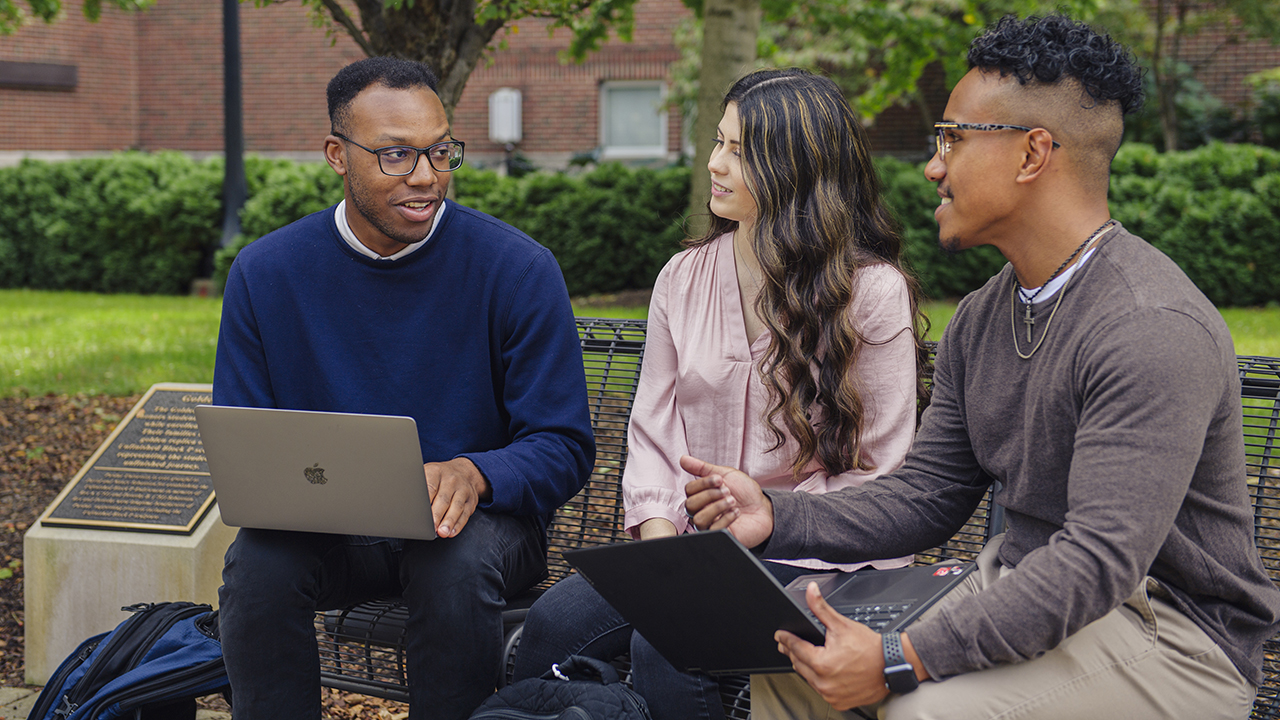 Three students work on laptops talking together sitting on a bench on campus