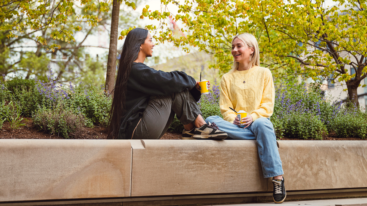 two students sit on a bench talking with coffee cups in hand