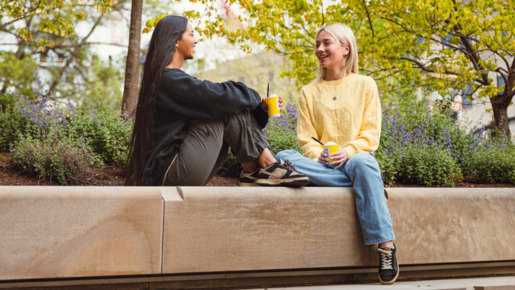 two students sit on a bench talking with coffee cups in hand