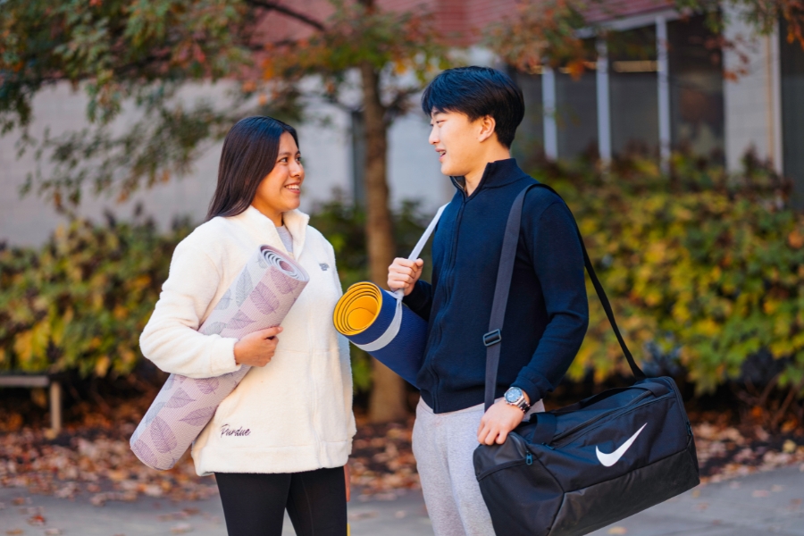 Two students carrying yoga mats and a gym bag chat outdoors on campus.