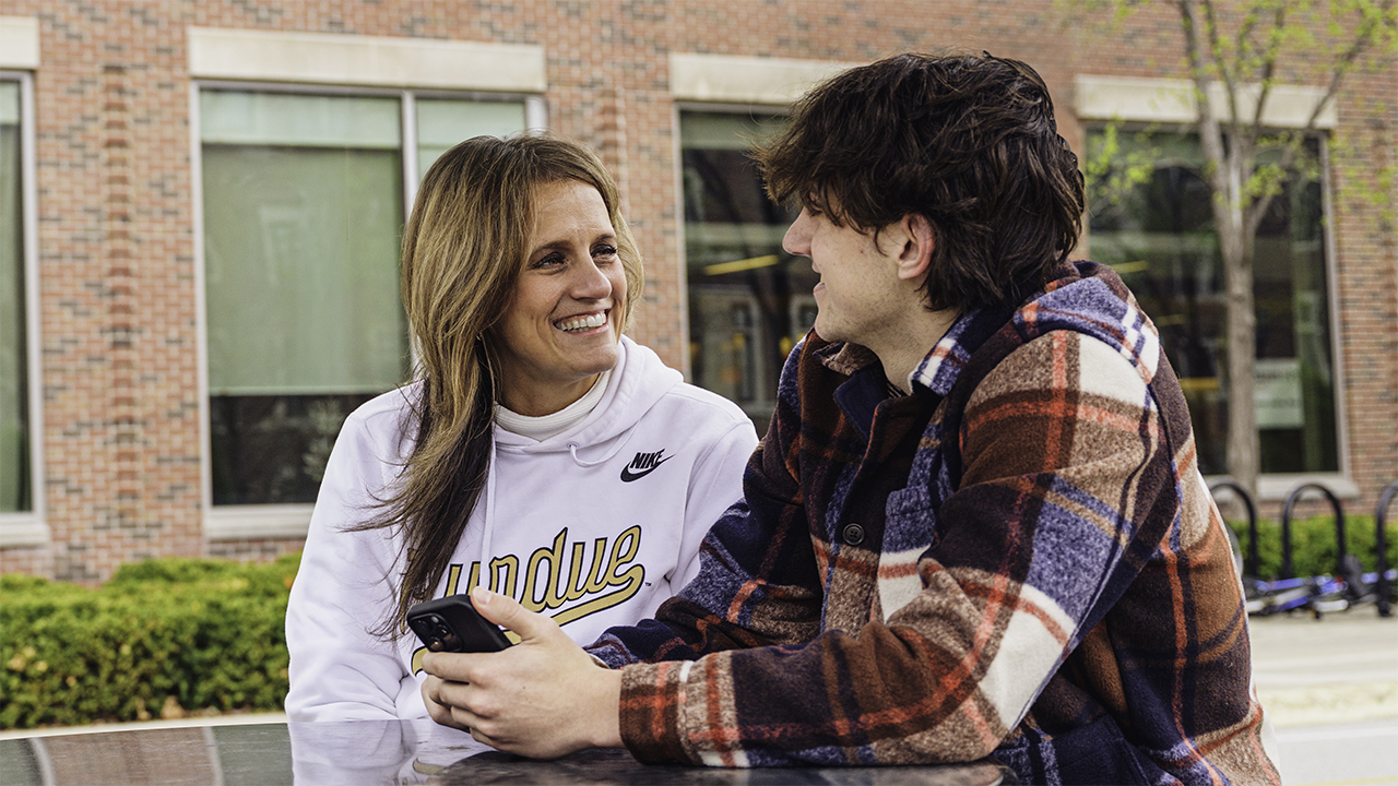 A parent sits with her student at an outdoor table while the student holds his phone