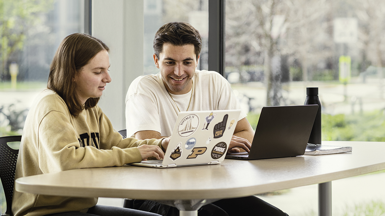Students sit together at a table with their laptops looking over one of the screens