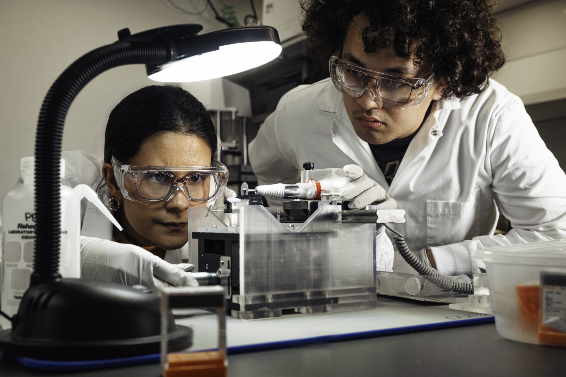Two students in lab coats and safety glasses closely examine equipment during a lab experiment.