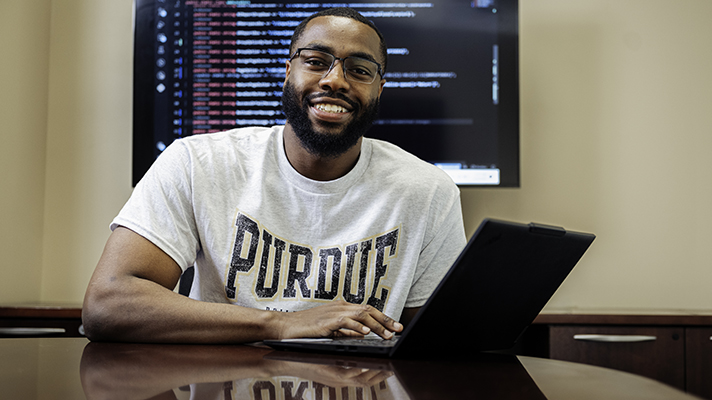 Prosper smiles as he looks up from his laptop with a large monitor displaying code on the wall behind him.