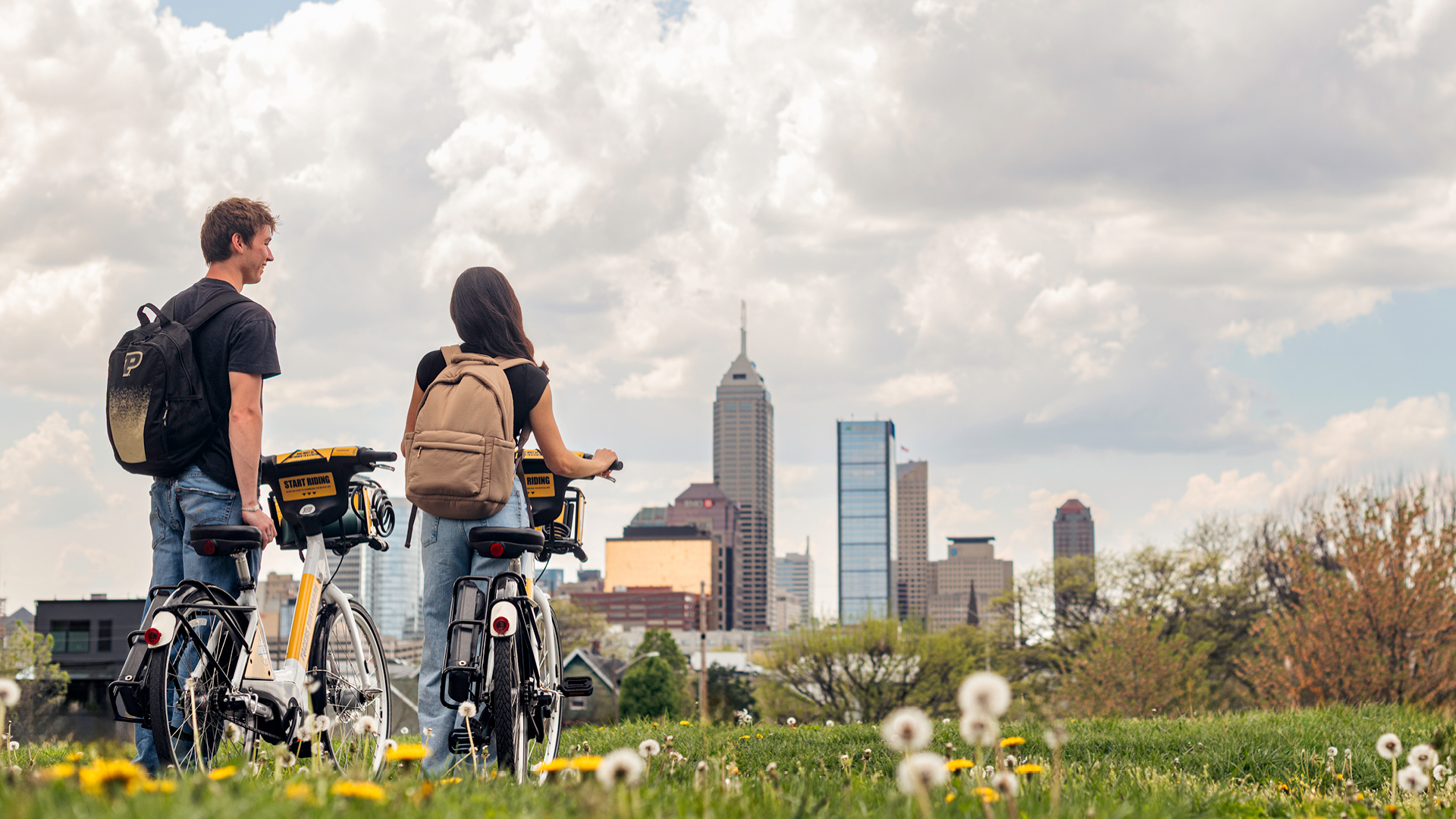 Two students with bike-share bicycles stand in a field overlooking the Indianapolis skyline.