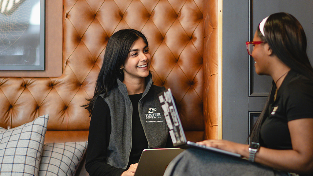 Two individuals engage in conversation while seated on a couch with a tufted leather backdrop, each using a laptop.