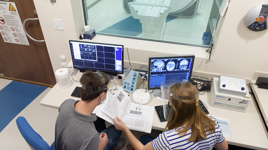 Two students reviewing a book with brain scan images on the computer screens in front of them
