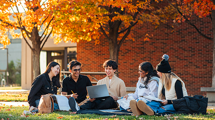 Five students sit on the ground on campus on a fall day, with notebooks and computers